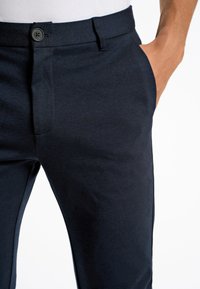 Marineblaue Hose mit glatter Textur, ausgestattet mit einer Vordertasche und einem Knopfverschluss. Leichter Stoff mit tailliertem Schnitt.