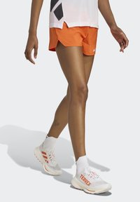 Oranžové atletické šortky s voľným strihom, v kombinácii s bielymi bežeckými topánkami, ktoré majú oranžové logo "TERREX" a farebné akcenty.