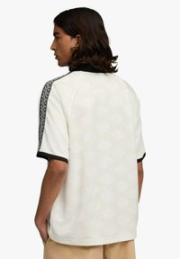 Polo a maniche corte in crema chiaro con motivo a diamante, colletto nero e dettagli laterali decorati. Vestibilità comoda con design semplice.