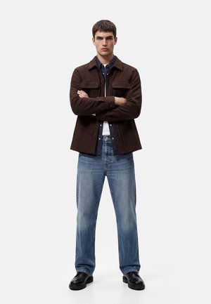 Joven de pie con los brazos cruzados, vestido con una chaqueta marrón oscuro, camisa azul marino, vaqueros azul claro y zapatos negros sobre fondo blanco.