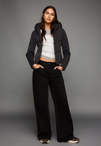 Giacca a vento nera con cappuccio, top corto grigio, jeans neri a gamba larga e sneaker scure. Texture lisce e design minimale.