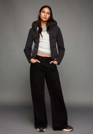 Črna puhovka s kapuco, sivi kratki top, široke črne jeans hlače in temni supergi. Gladke teksture in minimalni dizajn.