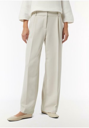 Frau trägt beige weite Hosen, cremefarbenen Pullover und passende beige Ballerinas, steht vor weißem Hintergrund.