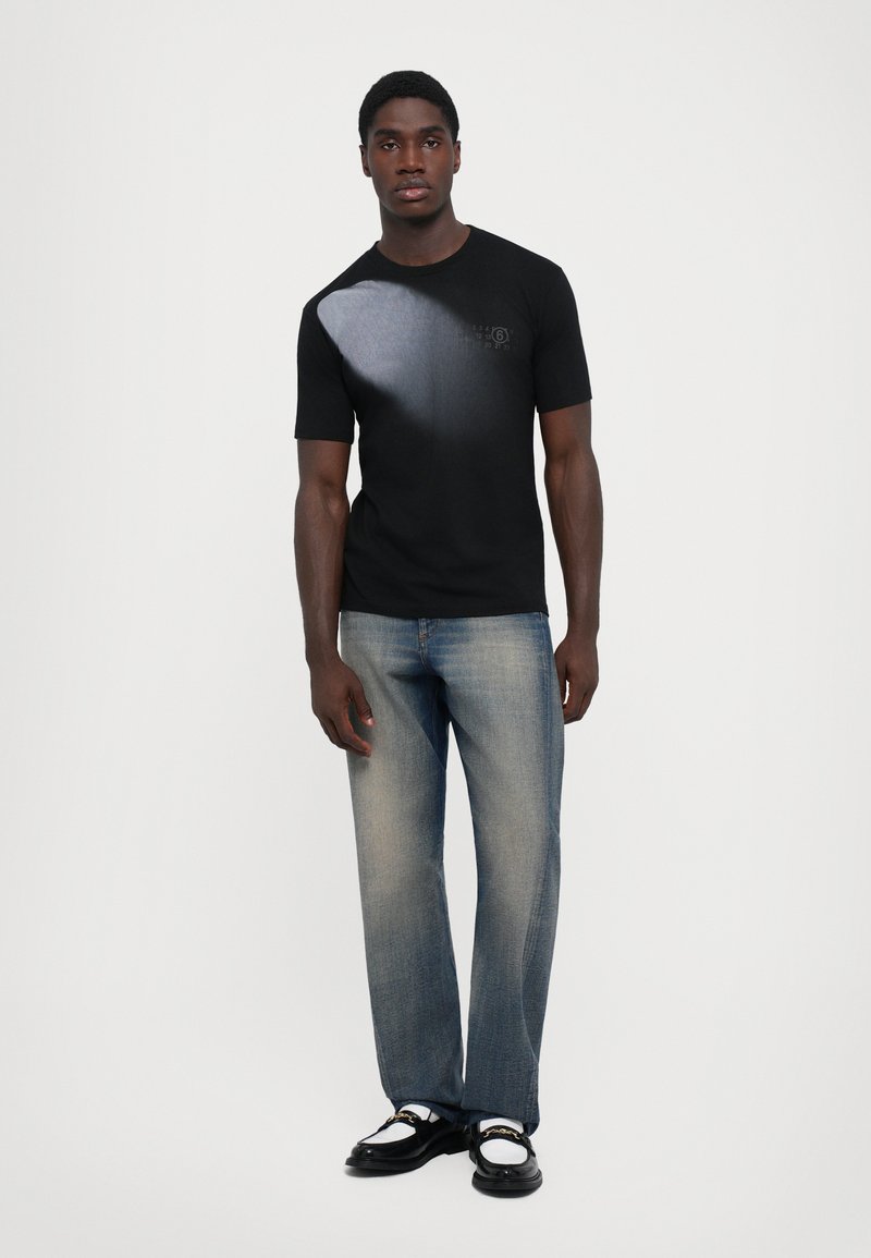 Ung man iklädd en svart graphic T-shirt, urtvättade raka jeans och svart-vita loafers, stående mot en enfärgad bakgrund.