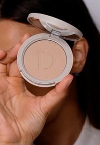 Polvo compacto redondo con un tono beige y un logo en relieve con la letra "D". La funda es de color gris claro y tiene una tapa transparente.