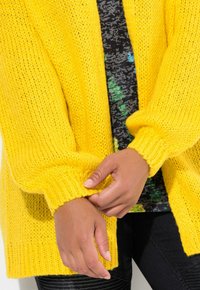 Cardigan giallo lavorato a maglia con una vestibilità morbida, polsini a coste e un motivo testurizzato, indossato sopra una maglietta stampata nera e verde.