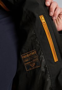 Čierna bunda s oranžovým zipsom, textilná etiketa s podrobnosťami o produkte a hladkou textúrou. Vyznačuje sa kontrastným zipsom.