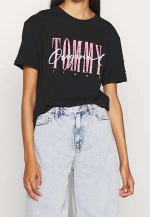 Woman wearing black Tommy Jeans t-shirt with pink floral letters and light blue high-waisted jeans, hands relaxed by sides.