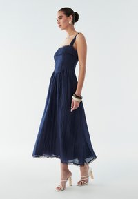 Vestido midi azul marino con tela texturizada, delicado encaje detallado y tirantes finos; combinado con tacones blancos de tiras y un brazalete grueso.