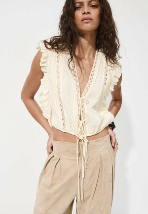 Femme aux longs cheveux bouclés portant un top sans manches crème à volants noué devant et un pantalon plissé beige taille haute.