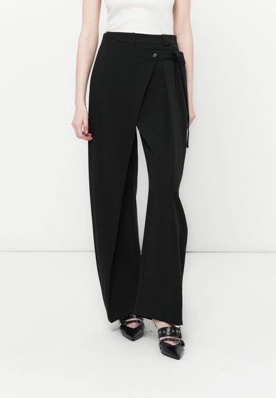 Femme portant un pantalon oversized noir avec des boutons et des détails noués, associé à des chaussures à talons noirs à bout pointu avec des brides.