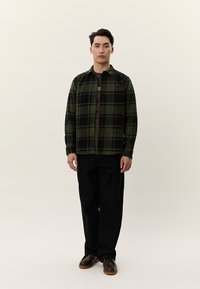 Jaunas vyras stovintis, dėvintis žalią ir juodą languotą marškinius ant tamsios marškinėlių, juodus kelnes ir rudus batus prieš paprastą foną.