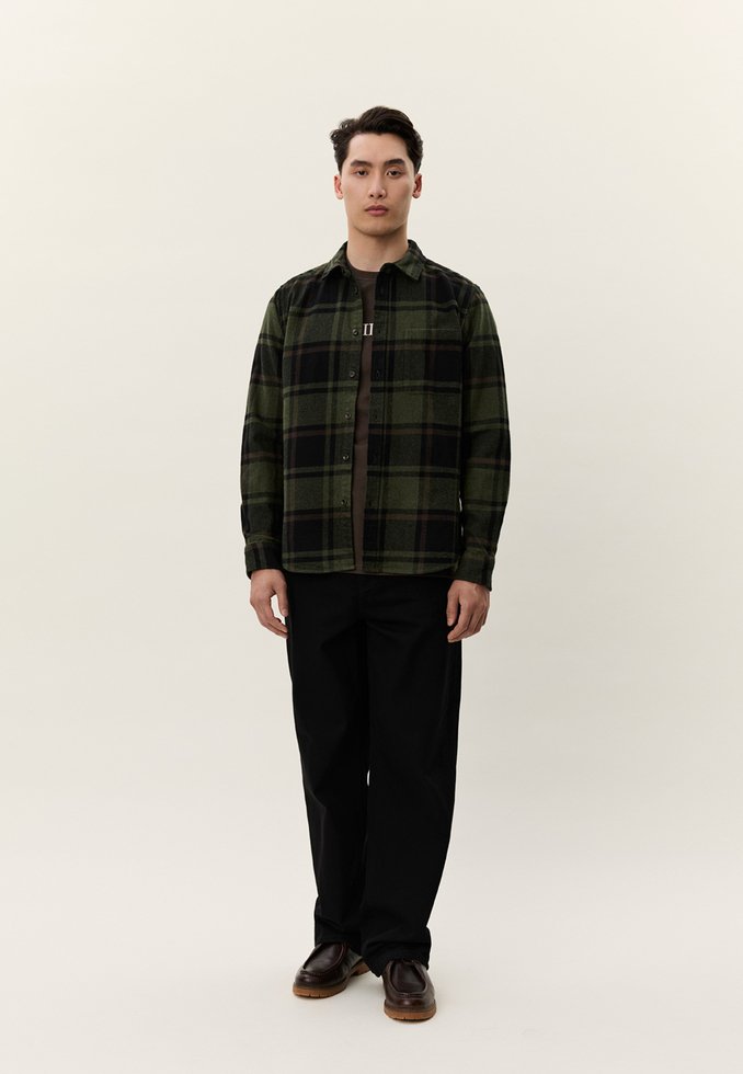 Jaunas vyras stovintis, dėvintis žalią ir juodą languotą marškinius ant tamsios marškinėlių, juodus kelnes ir rudus batus prieš paprastą foną.