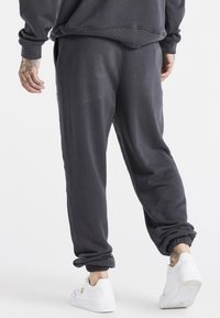 Man in een donkere grijze joggingbroek en witte sneakers, met getatoeëerde handen en enkels, staand tegen een eenvoudige witte achtergrond.