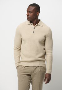 Beige gestrickter Pullover mit Kragen, drei Knöpfen und gerippter Textur, kombiniert mit hellbeigen Hosen.