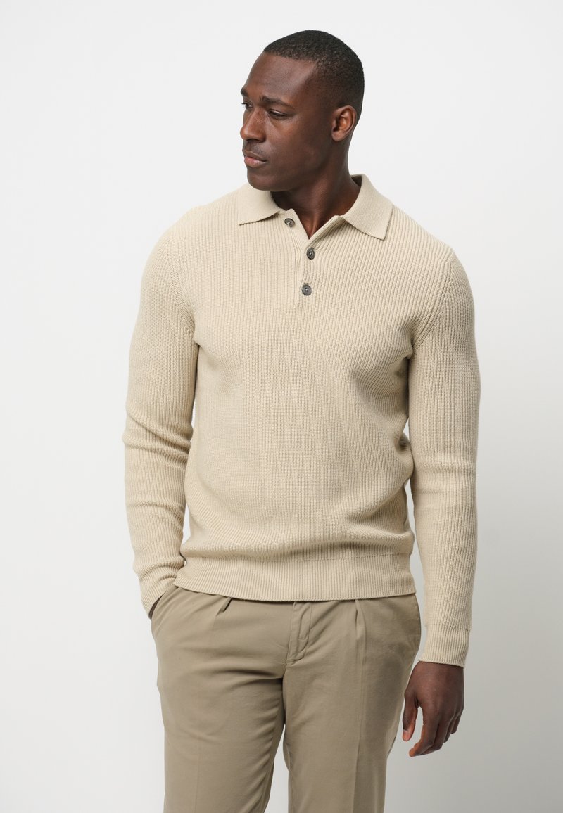 Beige gestrickter Pullover mit Kragen, drei Knöpfen und gerippter Textur, kombiniert mit hellbeigen Hosen.