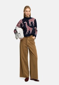 Granatowy sweter w wzór żyrafy w czerwono-białych kolorach; beżowe spodnie z szerokimi nogawkami; szara torebka z okrągłymi akcentami; bordowe buty.