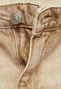 Bež denim tkanina s vidljivim patentnim zatvaračem i srebrnim gumbom izbliza. Teksturalna površina s detaljima šivanja uz rubove.