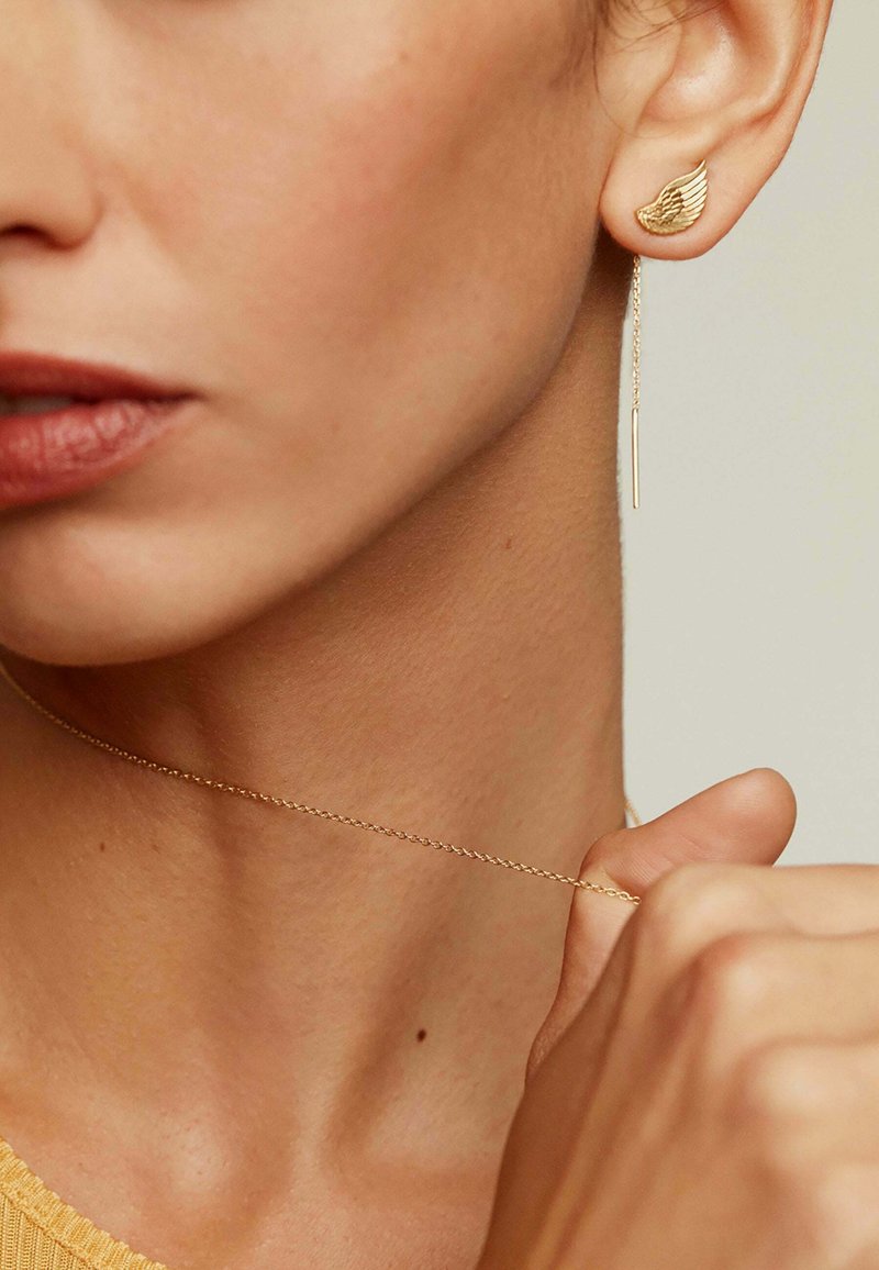 Pendientes de oro con un diseño de ala y un elemento de cadena colgante, exhibiendo una textura suave y un estilo minimalista. Piel visible en el fondo.