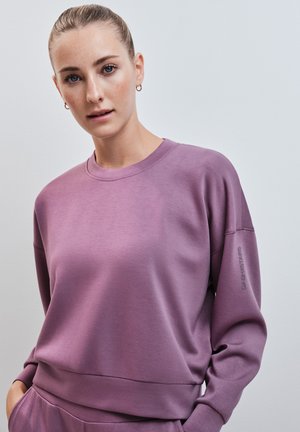 Jonge vrouw met blond haar in een knot, draagt een paarse trui met handen in de zakken van bijpassende paarse broek, tegen een effen achtergrond.