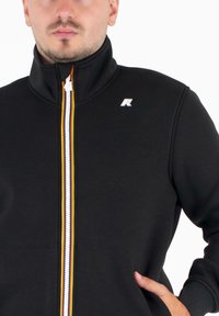 Zwarte zip-up sweatshirt met een hoge kraag, voorzien van een kleurrijke rits en een logo op de linkerschouder. Zacht, glad materiaal.