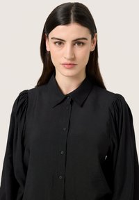 Camisa negra de botones con un cuello clásico, que presenta mangas abullonadas y un tejido suave y ligero.