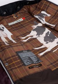 Barna kockás bélés, amelyen tehénillusztrációk találhatók, fekete ápolási címkével és egy címkével. Az anyag textúrássá és puhává tűnik. A cipzár széle látható.