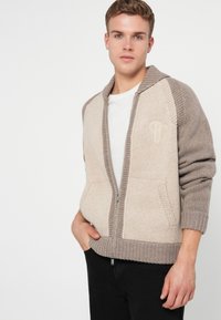 Jovem vestido com um hoodie de malha em bege e taupe com fecho, por cima de uma camisa branca, com calças pretas, de pé com uma mão no bolso.