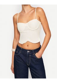 Bílý zkrácený top s kosticemi a volánkovým lemem. V kombinaci s tmavě modrými kalhotami s vysokým pasem. Hladká tkanina, tenké ramínka.
