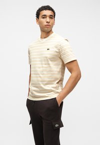 Νεαρός άνδρας με σγουρά μαλλιά φοράει μπλουζάκι με μπεζ και λευκές ρίγες και μαύρο cargo παντελόνι με λογότυπο της μάρκας στο στήθος και στην τσέπη, με τα χέρια στις τσέπες.