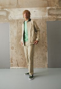 Beige Baumwolljacke mit schwarzem Kragen, grüner Sweatshirt, beige Hose mit sichtbaren Nähten und schwarzen Schuhen mit grünem Muster.