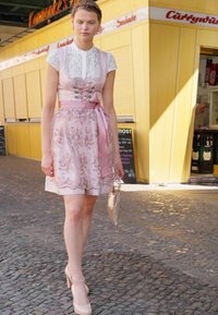 Robe rose avec des détails en dentelle, un corsage ajusté et une jupe fluide, accentuée par une large ceinture en ruban. Portée avec des talons beiges et une pochette.