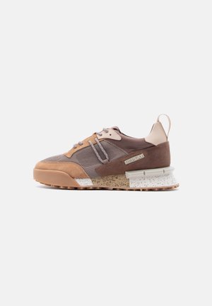 Brown and tan sneaker with suede and mesh material, textured accents, reflective laces, and a speckled outsole, featuring a prominent logo.