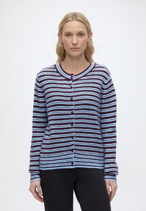 Femme aux cheveux blonds portant un cardigan rayé bleu et bordeaux boutonné et un pantalon noir, debout devant un fond clair uni.
