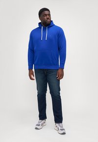 Hanorac albastru cu snururi și buzunar frontal, asociat cu blugi închisi și adidași deschiși. Design simplu și casual; textură netedă.