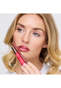 Deux crayons à lèvres, un rouge foncé et un marron clair, tenus à côté d'une lèvre de teinte neutre. Texture lisse et mate avec un packaging élégant.