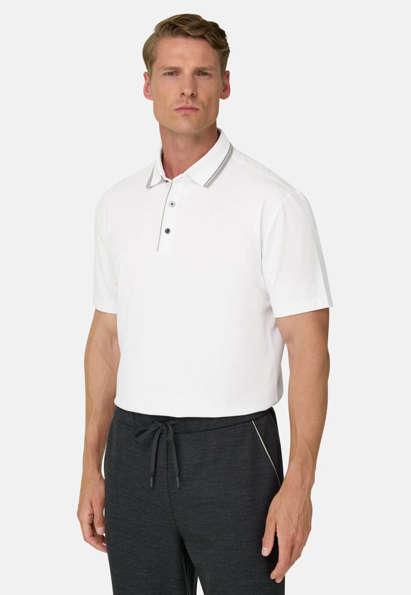 Homme portant un polo blanc à manches courtes avec des bordures grises et un pantalon à cordon gris foncé sur un fond clair.