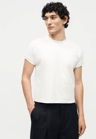 Νέος άνδρας με κατσαρά μαλλιά, φορώντας λευκό κοντομάνικο μπλουζάκι και μαύρο παντελόνι, κοιτάζει προς τα πλάγια με το ένα χέρι στην τσέπη.