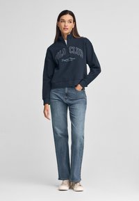 Felpa cropped blu marina con la scritta "POLO CLUB" stampata in bianco, abbinata a jeans azzurri a gamba dritta e sneakers color crema.