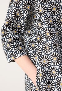 Bras avec la main dans la poche, portant une chemise recouverte de motifs géométriques circulaires noirs et blancs avec des centres jaunes et des contours bleus.