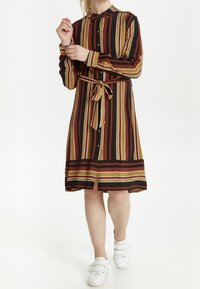 Abito camicia a righe in nero, marrone e senape, con colletto, bottoni e vita legata. Presenta un taglio al ginocchio e uno spacco laterale.