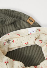 Two fabric pieces: a textured olive green piece and a cream-colored one with a small red cherry pattern, featuring a leather tag sewn on.