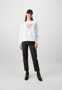 Biela mikina s červeným trojuholníkovým logom a nápisom "GUESS?" kombinovaná s čiernymi džínmi a čiernymi členkovými čižmami. Nosená čierna šiltovka.