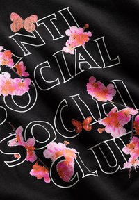 Czarny materiał zawiera napis "ANTI SOCIAL CLUB" w pogrubionych białych literach, ozdobiony różowymi kwiatami i pomarańczowymi motylami.