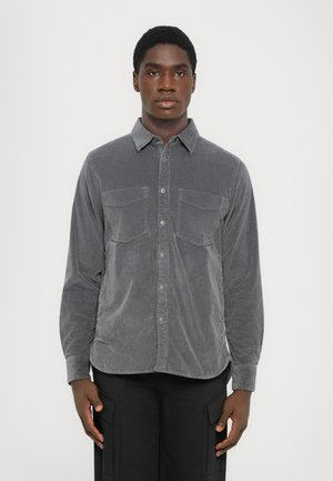 Young man standing facing forward wearing a grey corduroy button-up shirt and black pants with large cargo pockets.