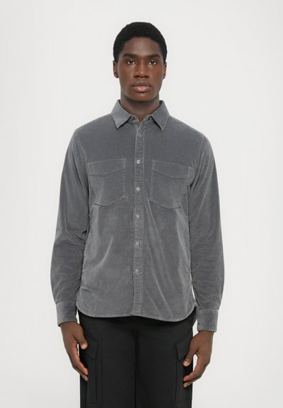 Young man standing facing forward wearing a grey corduroy button-up shirt and black pants with large cargo pockets.