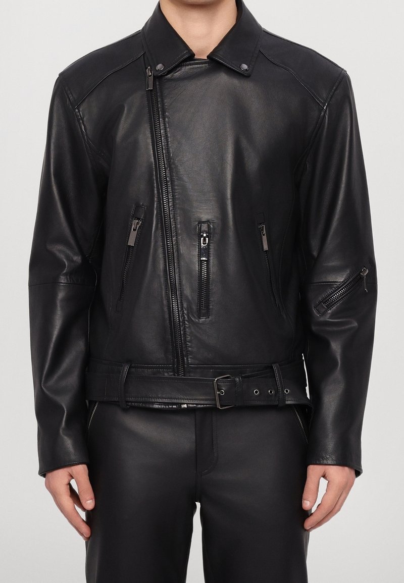 Veste en cuir noire asymétrique à fermeture éclair avec poches zippées sur les côtés et les manches et ceinture à la taille, portée par une personne.