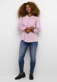 Blusa rosa a maniche lunghe con motivo testurizzato, chiusura a bottoni e dettagli in pizzo sui polsini, abbinata a jeans skinny blu e mocassini neri.
