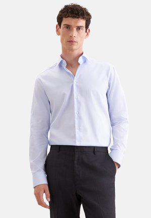 Junger Mann mit lockigem Haar, der ein hellblau gestreiftes Langarmhemd und schwarze Hose trägt, steht mit einer Hand in der Tasche.