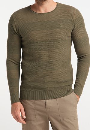 Mężczyzna w oliwkowozielonym teksturowanym swetrze z długim rękawem i beżowych spodniach, z ręką w kieszeni, na tle jednolitego białego tła.